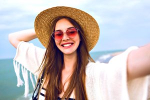 Jonge gelukkige blonde meid die selfie maakt met een schattige zonnebril met strohoed bij de zee